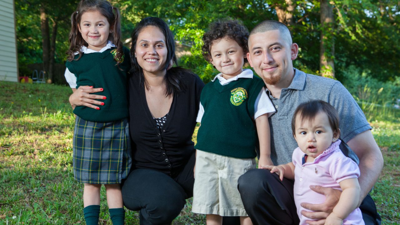 Two parents squatting with their three young children in front of wooded residential yard
