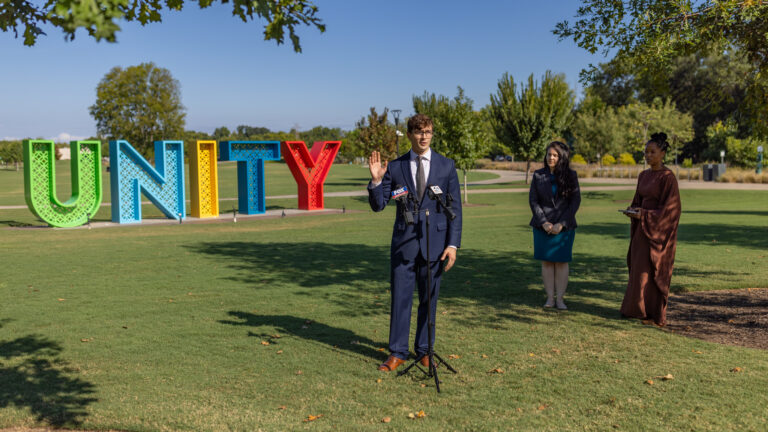 Man speaks at microphones in park, large UNITY sculpture and two women visible behind him