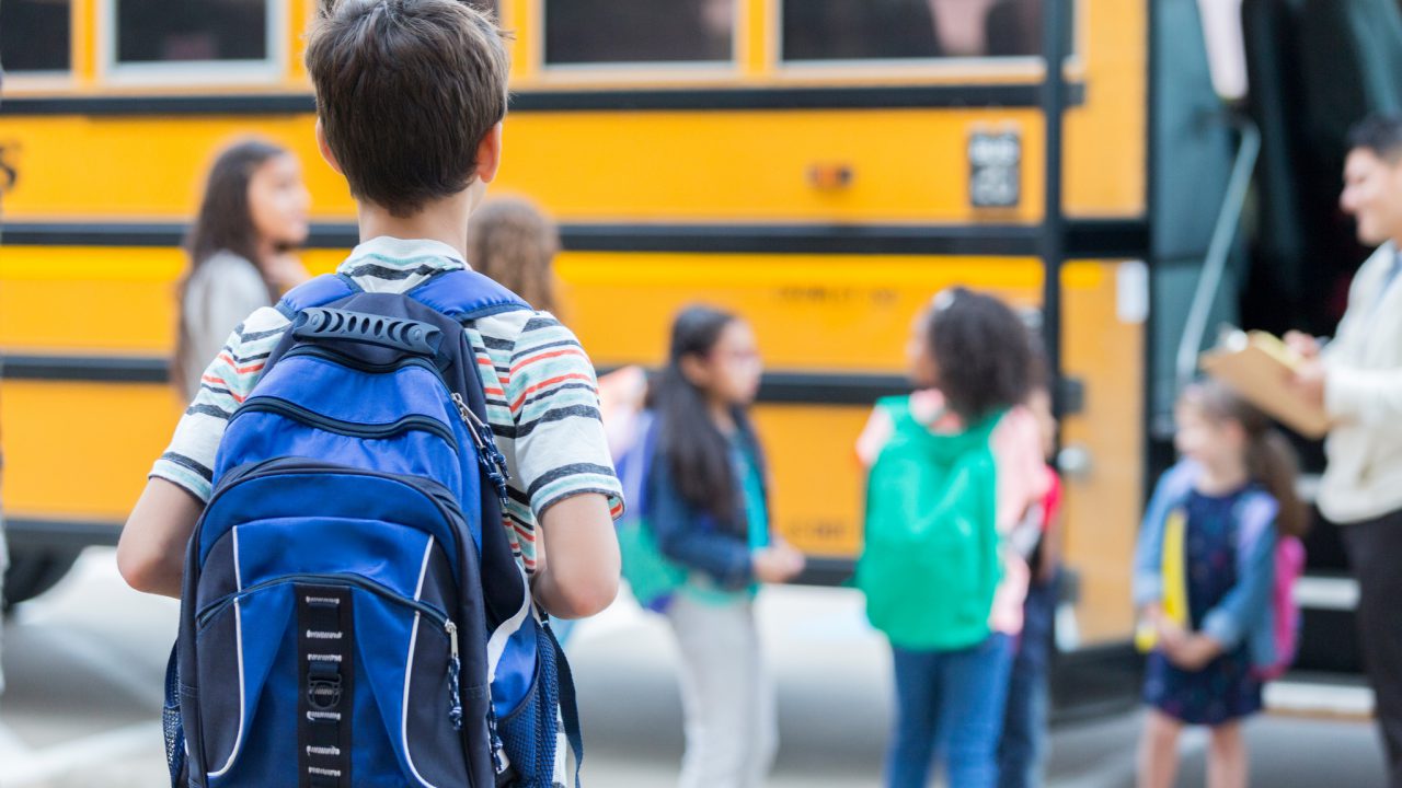 Rear view of elementary age boy waiting to get on school bus. His classmates are loading the bus in the background.