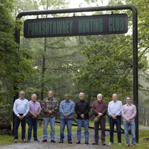 Eight men stand in a row beneath a wooden sign, surrounded by trees at a forested property