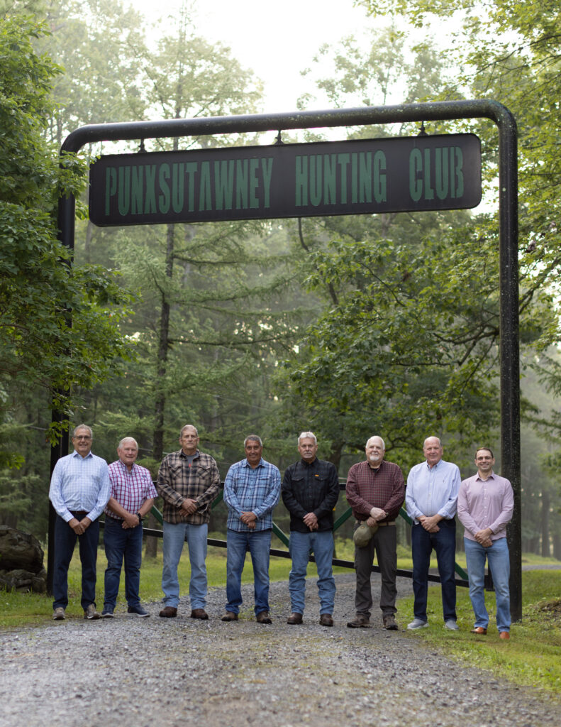 Eight men stand in a row beneath a wooden sign, surrounded by trees at a forested property