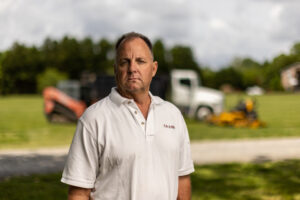 man stands in front of lawn care equipment and dump truck