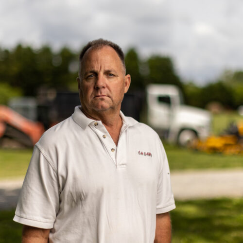 man stands in front of lawn care equipment and dump truck