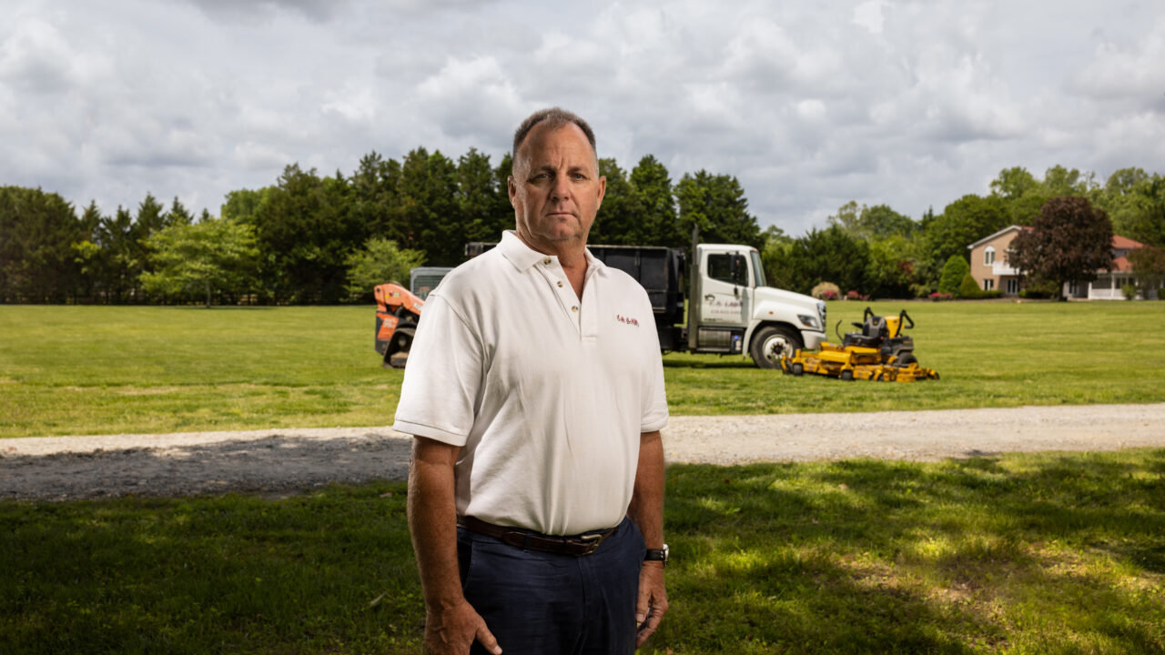 man stands in front of lawn care equipment and dump truck
