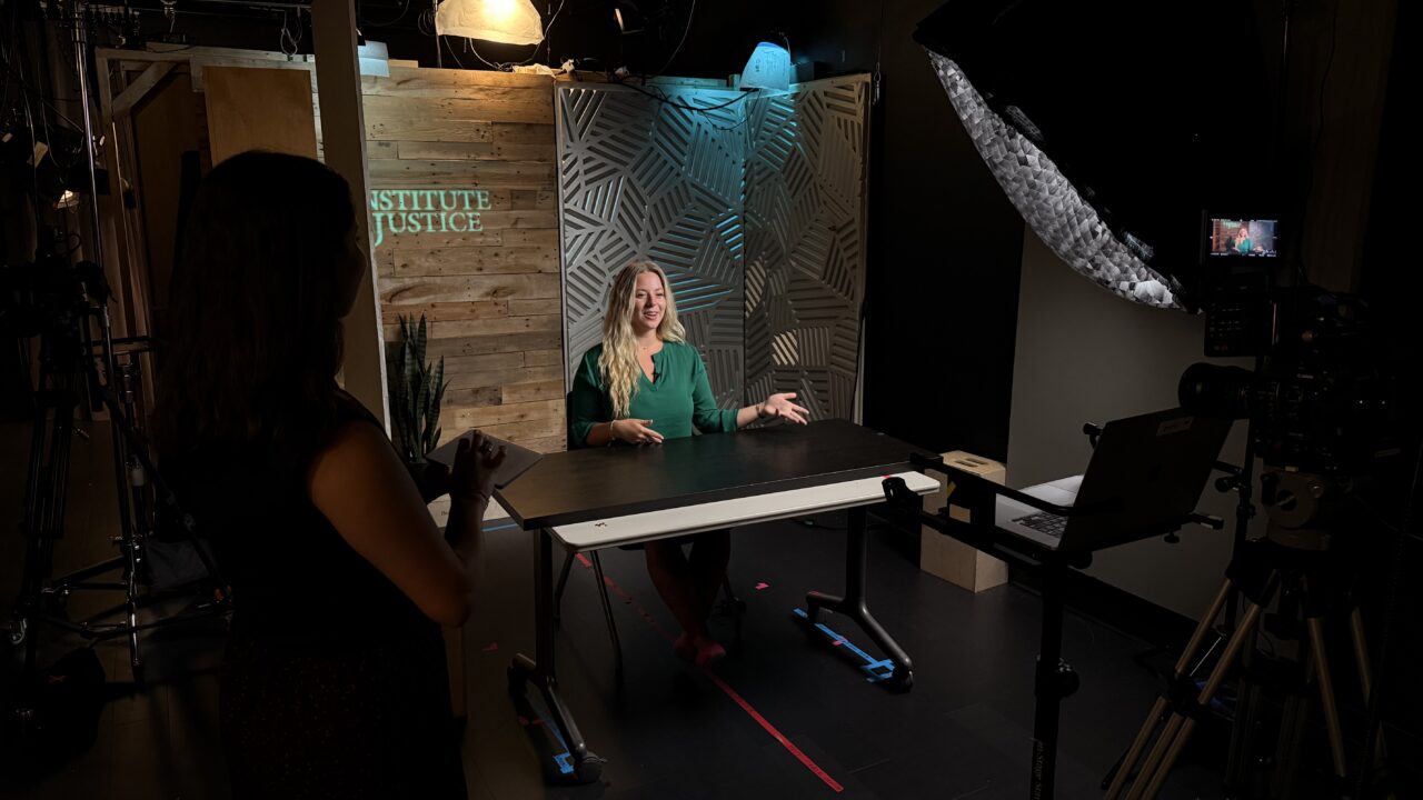 Woman in green shirt interviewed at a table in a TV studio with Institute for Justice backdrop