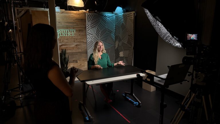 Woman in green shirt interviewed at a table in a TV studio with Institute for Justice backdrop