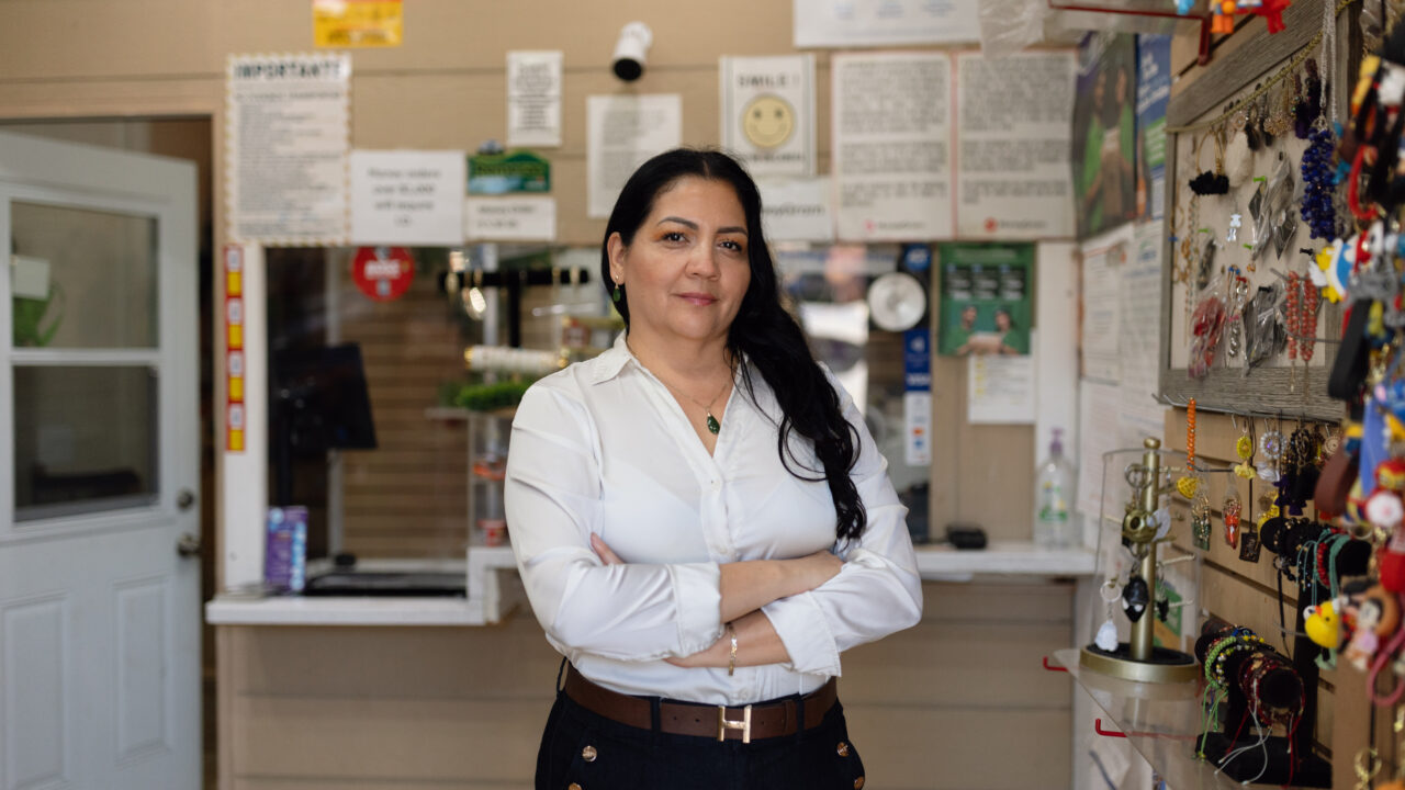 Esperanza Gomez stands with arms crossed in her shop, jewelry display behind her