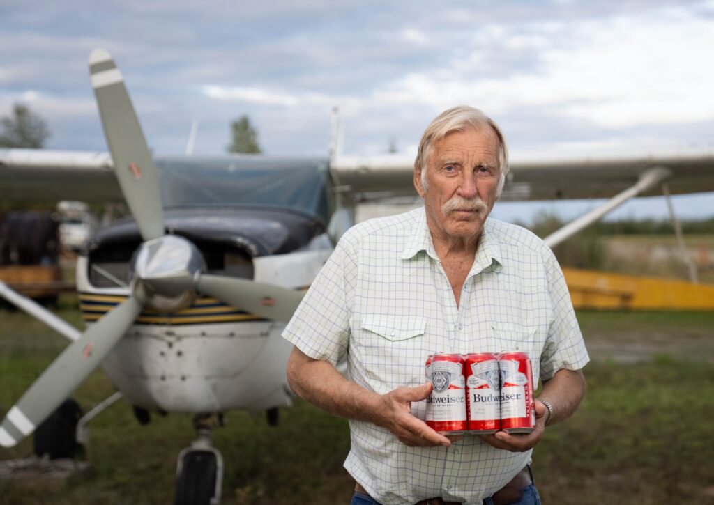 Ken Jouppi holds a six-pack of Budweiser beside a small plane on a grassy airstrip