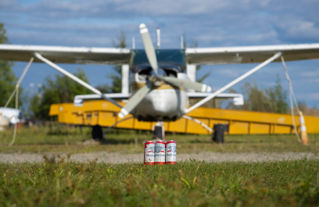 Cans of Budweiser on grass in front of a small crop-duster plane, referencing an Alaska plane seizure