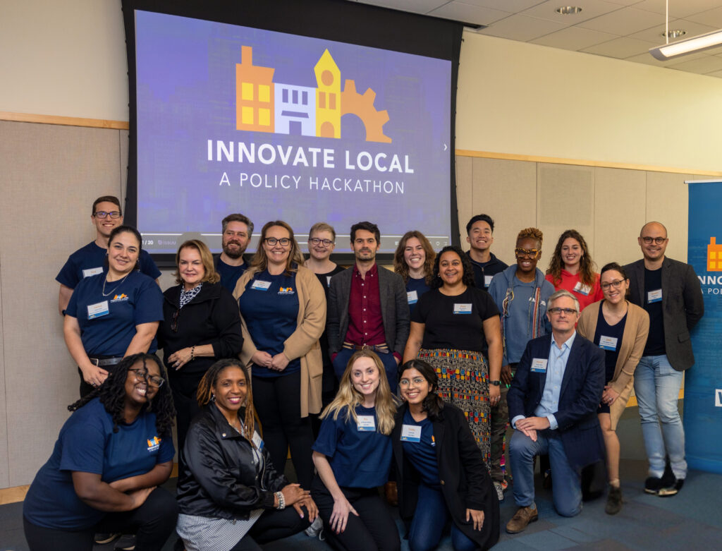 Group poses beneath Innovate Local: A Policy Hackathon screen, wearing name tags and event T-shirts.