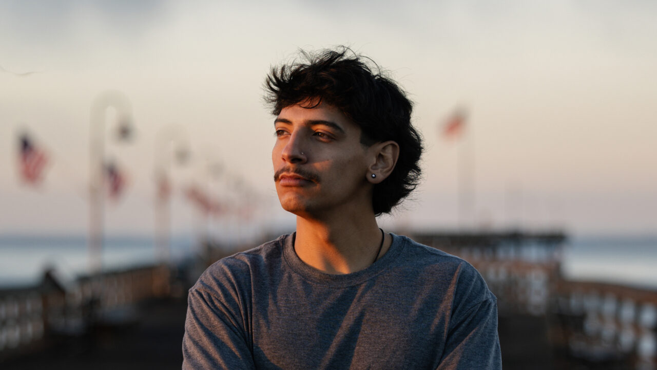 George Retes, wearing a gray t-shirt with an earring, looks into the distance outdoors near the ocean at dusk