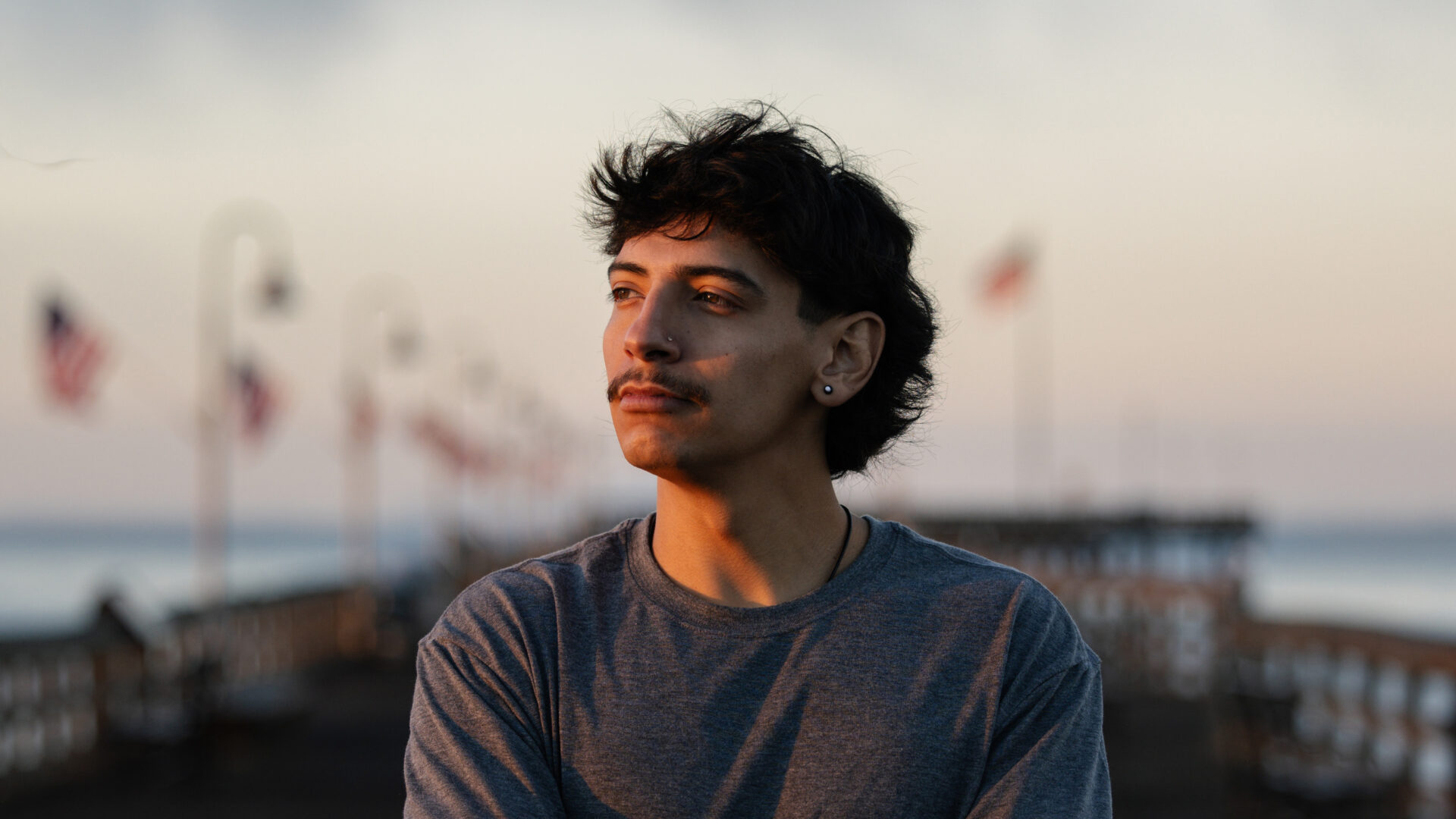 George Retes, wearing a gray t-shirt with an earring, looks into the distance outdoors near the ocean at dusk