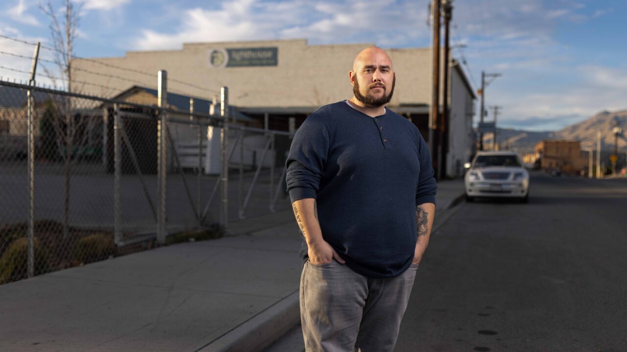 Bearded man with tattoos stands on sidewalk near chain-link fence and city street