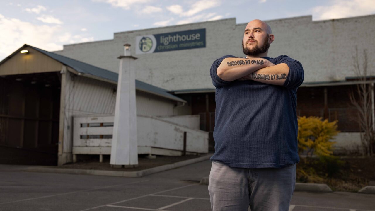 Bald man with arm tattoos reading Rescued by Grace stands outside Lighthouse Christian Ministries building