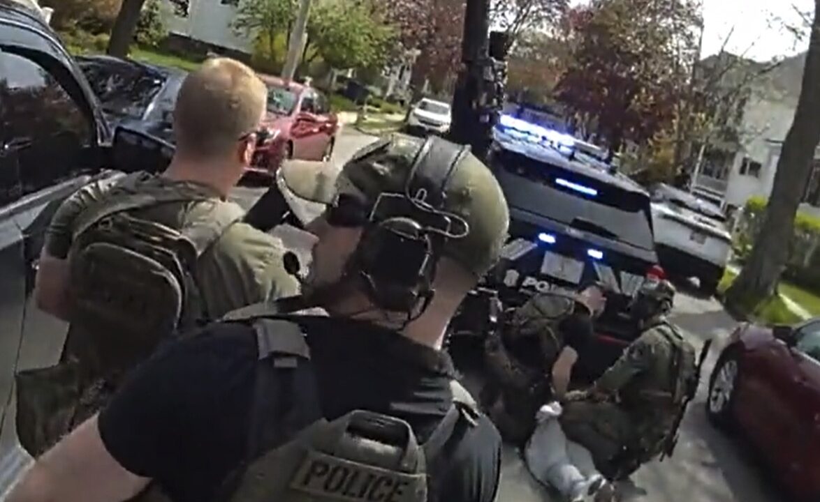 Body camera footage showing multiple officers in tactical gear surrounding a person near vehicles on a residential street