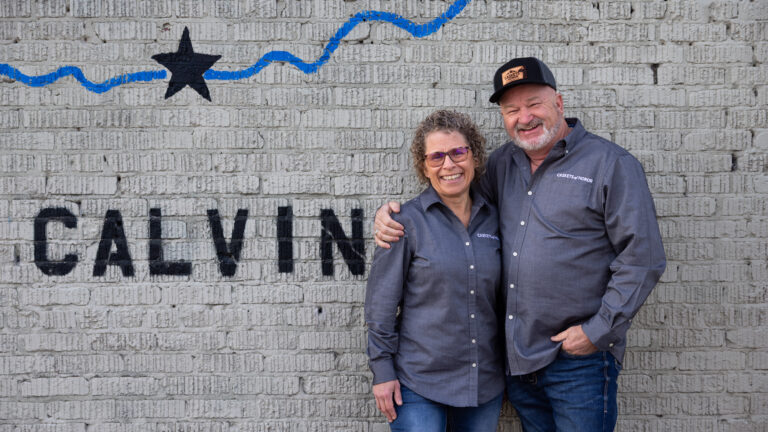 A woman and man in matching gray work shirts stand together in front of a painted brick wall reading 'Calvin' with a blue wavy line and star.