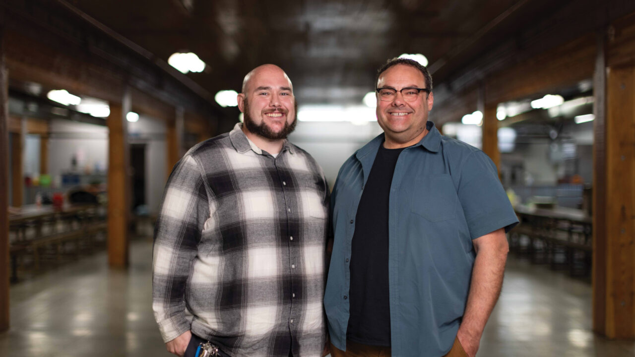 Two men standing in industrial warehouse space with overhead lighting and workbenches visible in background