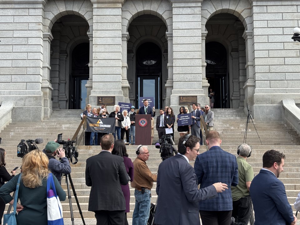 Press conference on stone steps of government building with speaker at podium surrounded by supporters facing reporters and cameras