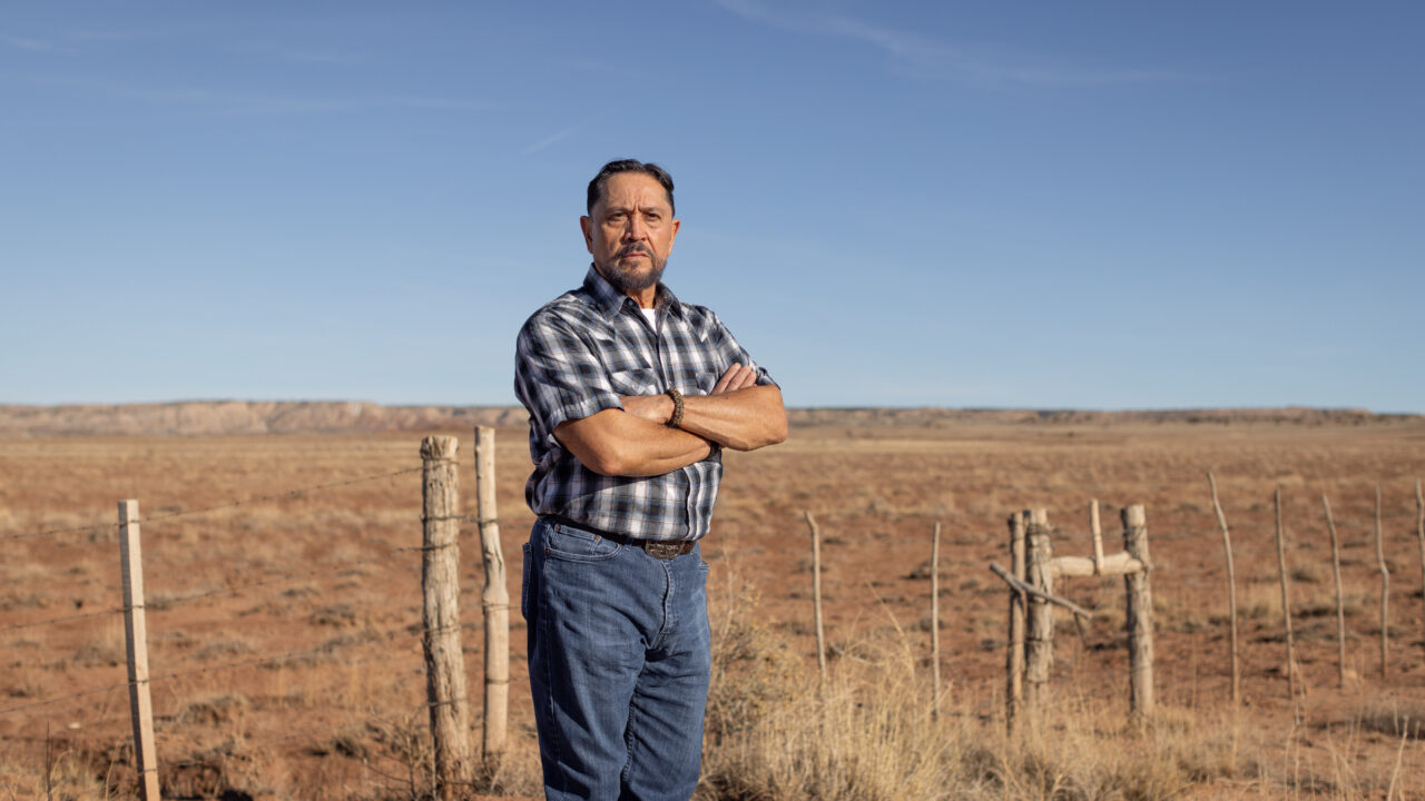 Fernie Madrid stands with crossed arms in an arid landscape with weathered fence posts and sparse vegetation under clear blue sky