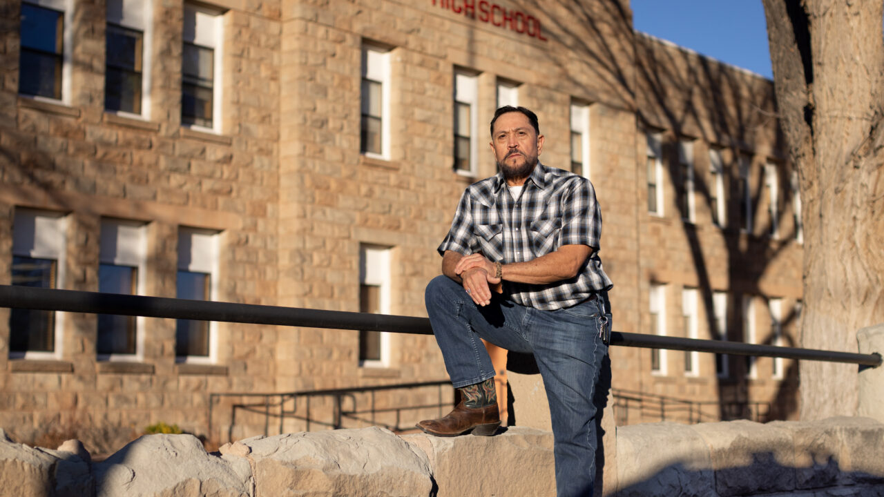 Educator Fernie Madrid stands outside a brick school building with a black metal railing in the foreground