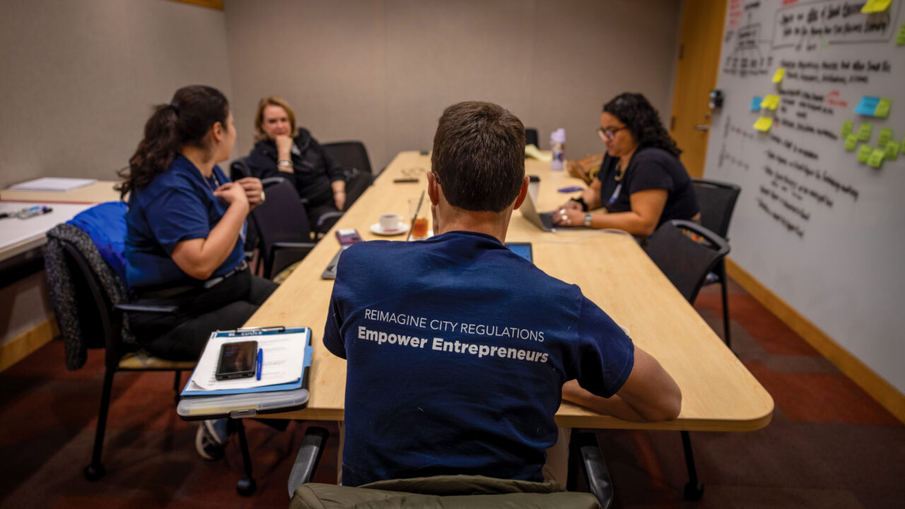 Five people in a conference room meeting, person facing away wears navy shirt reading 'REIMAGINE CITY REGULATIONS Empower Entrepreneurs