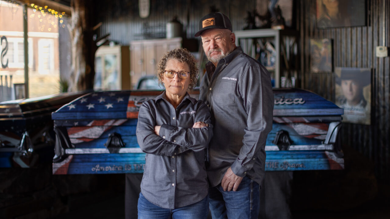 Woman and man in matching work shirts standing in workshop in front of casket with American flag design