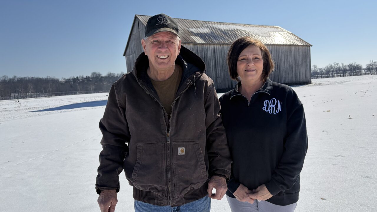 David and Debbie Ross standing in a snow-covered field with a weathered barn behind them on their Triple R Farms property