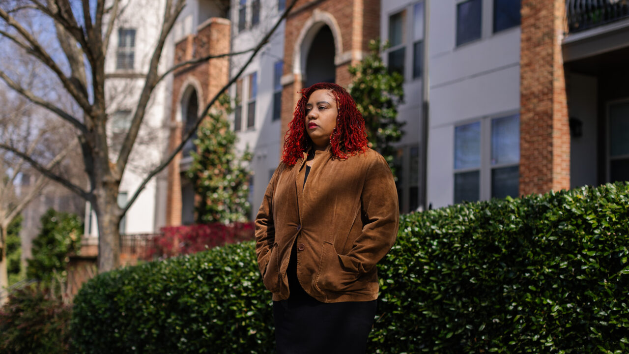 Cathy George stands outside a residential building with brick and arched windows, wearing a brown jacket.