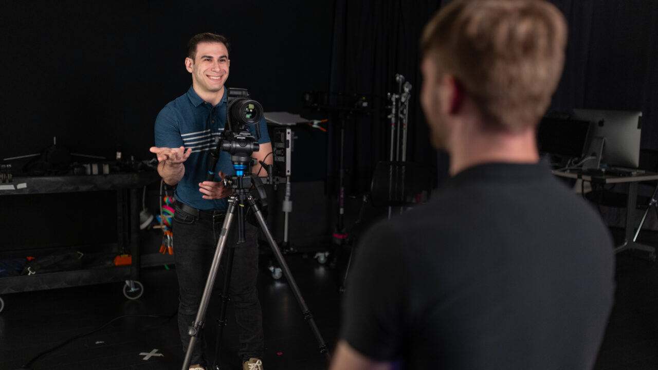 Man in blue polo shirt gesturing while standing behind video camera on tripod in studio, with another person in foreground