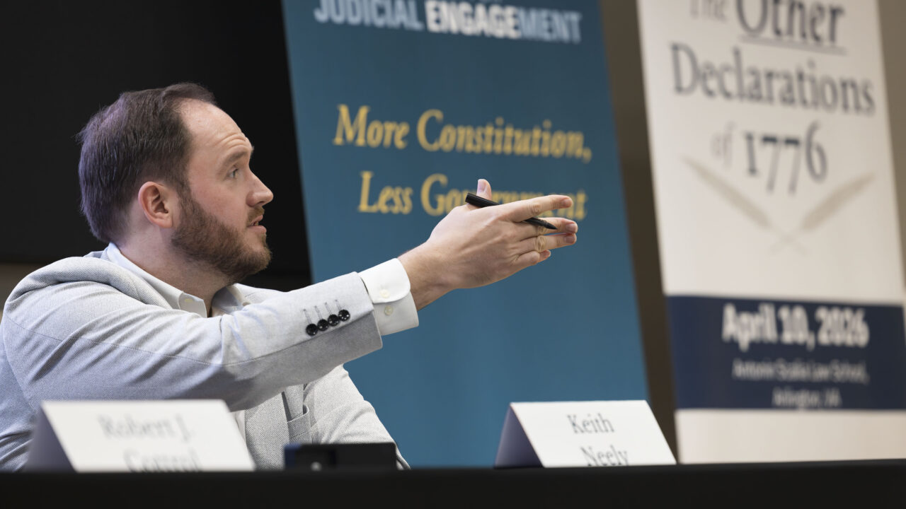 Man in light gray suit jacket gestures with pen during panel discussion, seated at table with name placard and banners about Constitution and