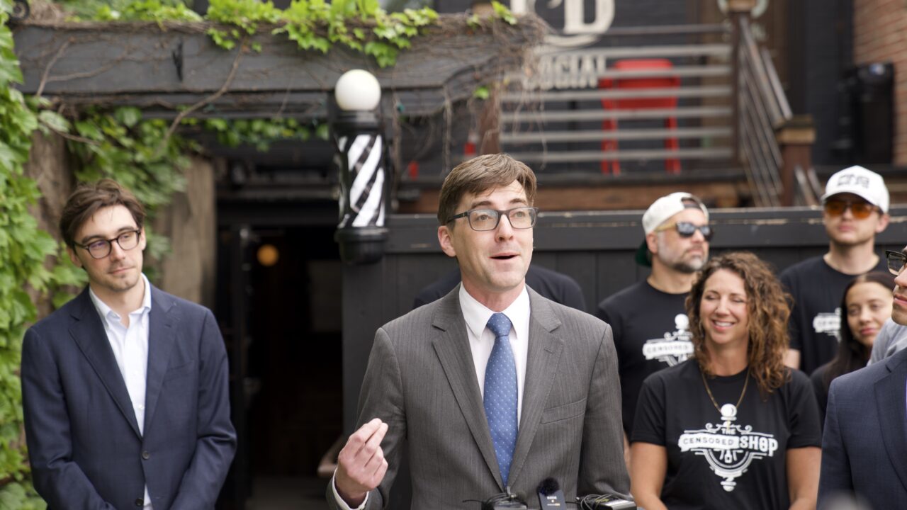 Man in gray suit and blue tie speaks outdoors, flanked by people in black t-shirts with barbershop logos and others in business attire