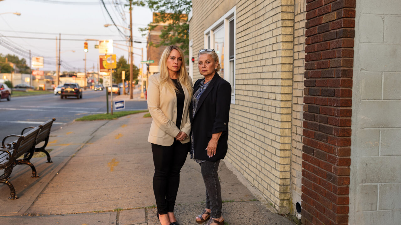 Honey Meerzon and Dina Finkelstein stand on a sidewalk beside their apartment building in an urban neighborhood at dusk.