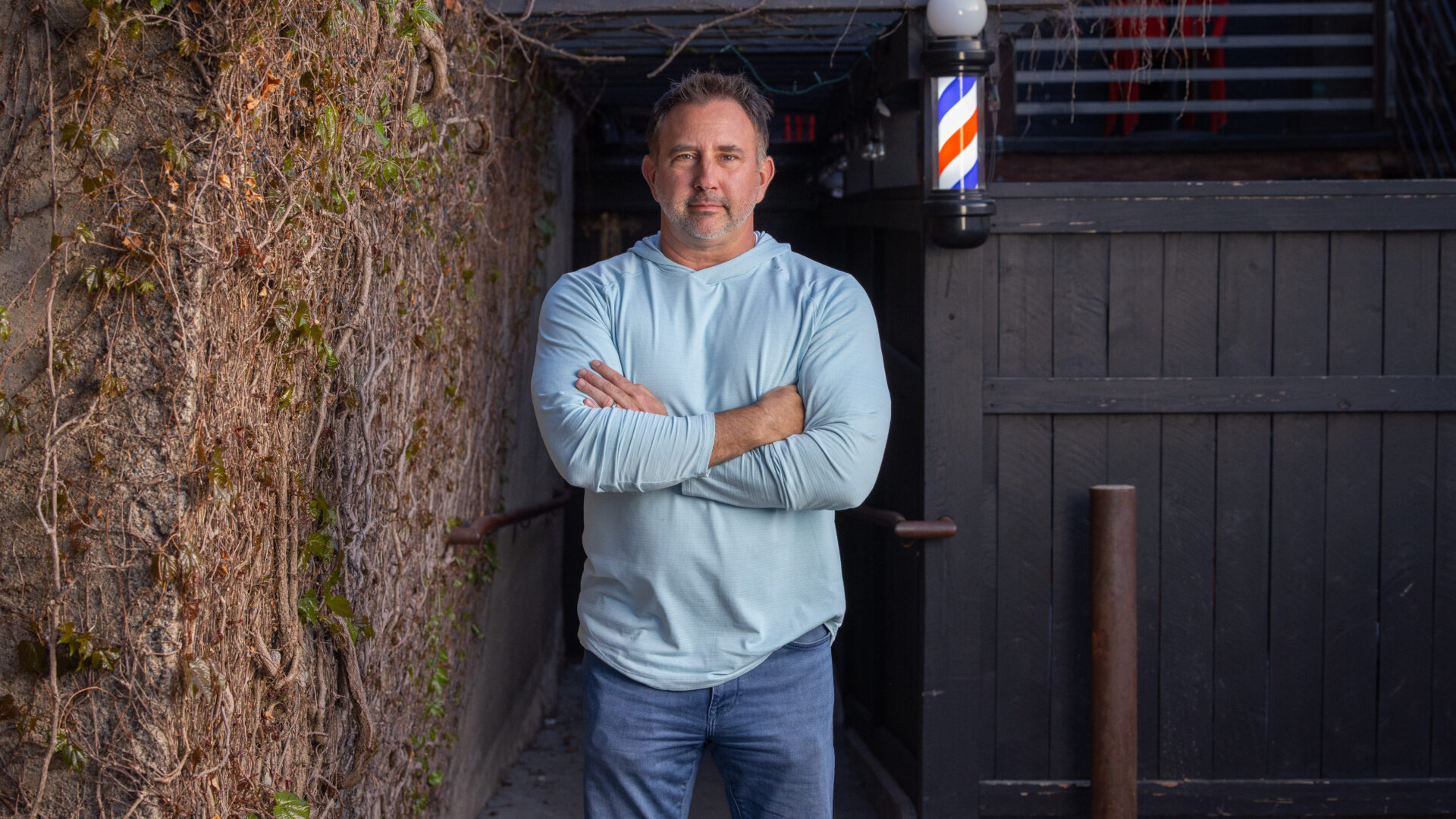 Man with crossed arms stands in front of a barbershop entrance with a traditional barber pole visible inside.