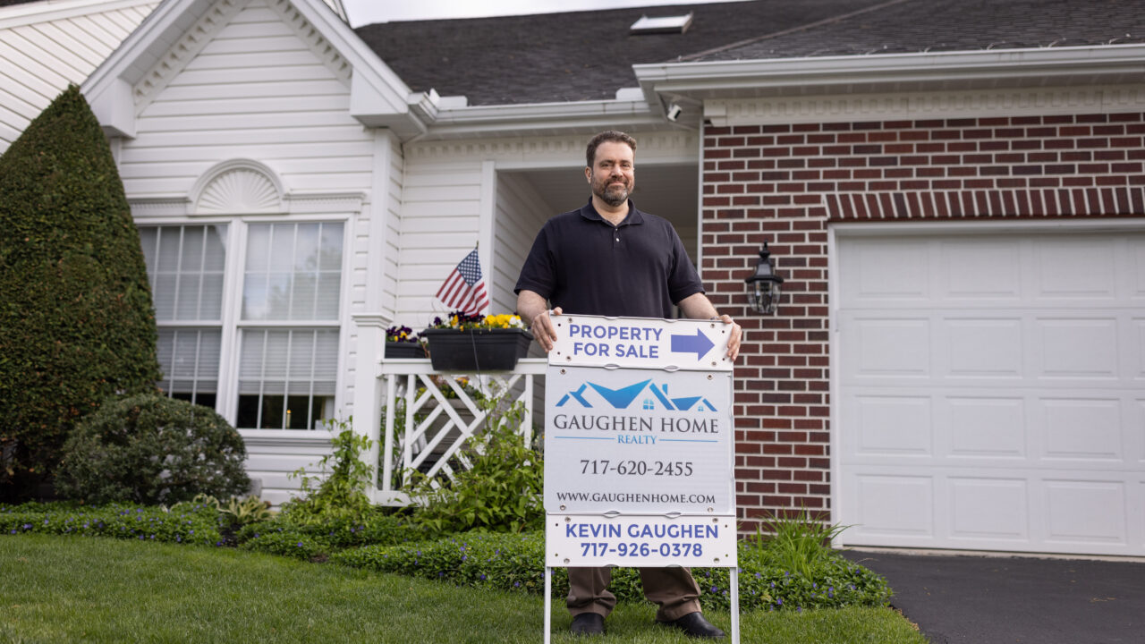 Kevin Gaughen in navy polo stands behind Gaughen Home Realty sign reading 'Property For Sale' on residential lawn with white siding and brick home.