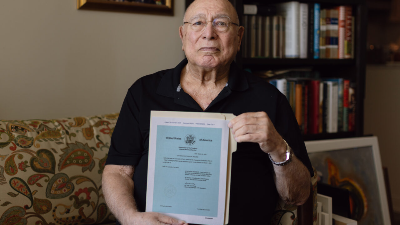 Man wearing glasses holds up a U.S. Department of the Treasury document with official seal while seated on a couch with bookshelves behind him.