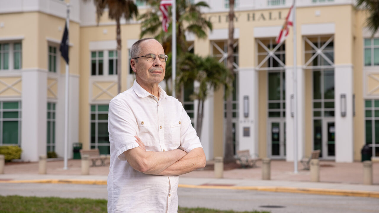 Man with arms crossed wearing glasses and white shirt standing in front of North Port City Hall with palm trees