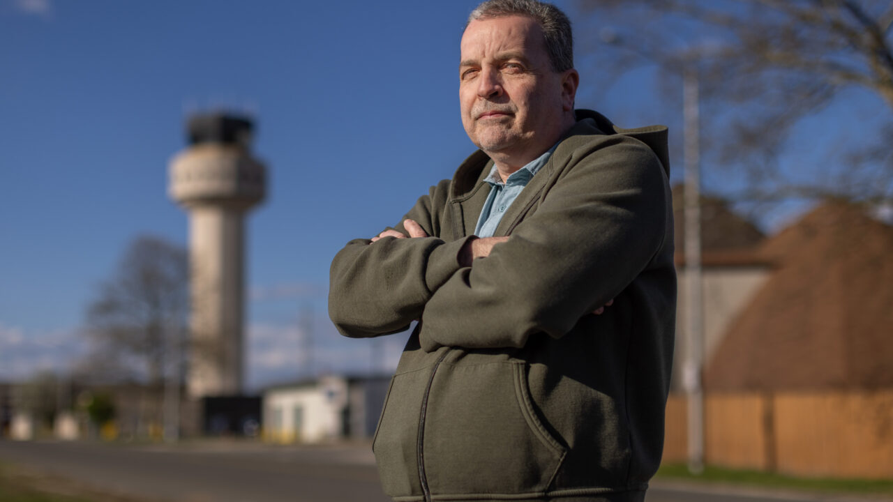 Man with arms crossed wearing olive hoodie stands outdoors with airport control tower visible in blurred background.