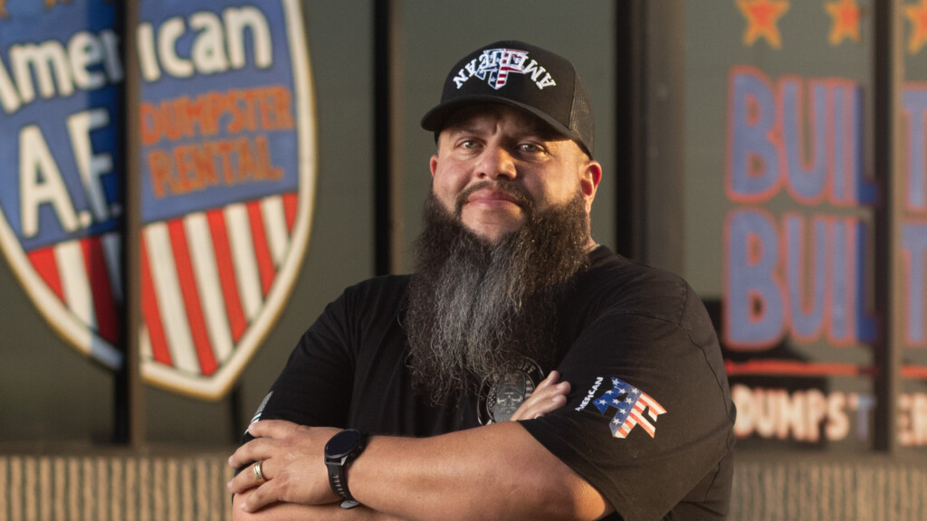 Man with long beard stands with arms crossed wearing black t-shirt with American flag patch, corrugated metal wall behind him.
