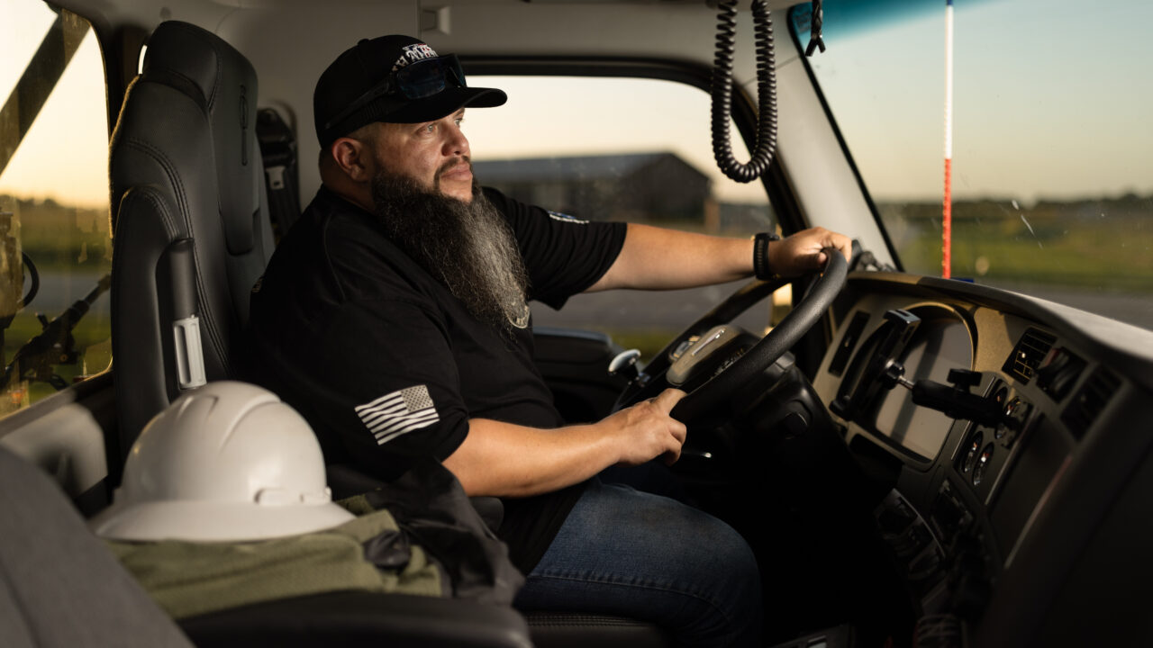 Man with beard and cap seated in driver's seat of large commercial truck, hands on steering wheel, with white hard hat visible on passenger seat.