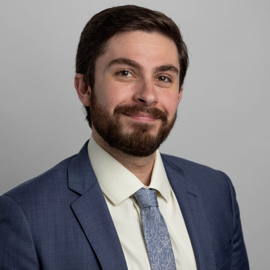 Man with beard wearing navy blazer, white dress shirt, and patterned tie against gray background