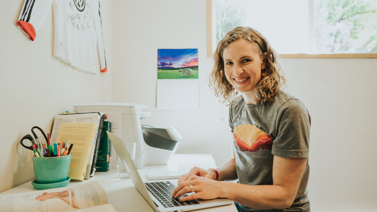 Woman with curly hair smiling at laptop on white desk with green pen holder, open book, and children's artwork on wall