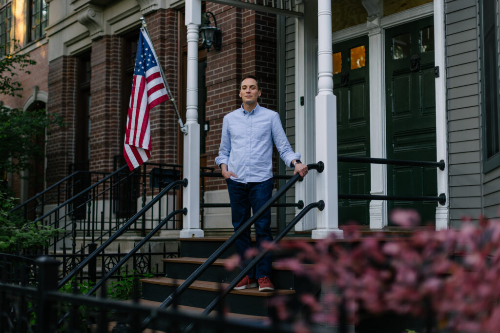 Man in light blue shirt standing on front porch steps beside American flag and green shutters