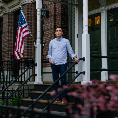 Man in light blue shirt standing on front porch steps beside American flag and green shutters