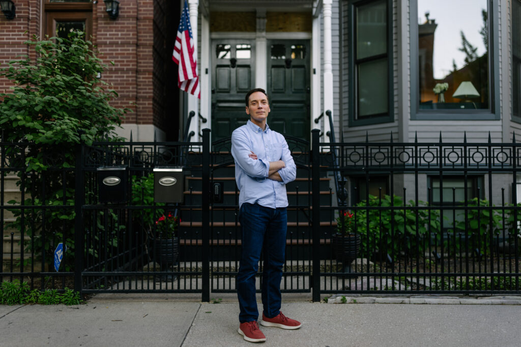 Man with crossed arms standing on sidewalk in front of brick building with green door, decorative black iron fence, and American flag