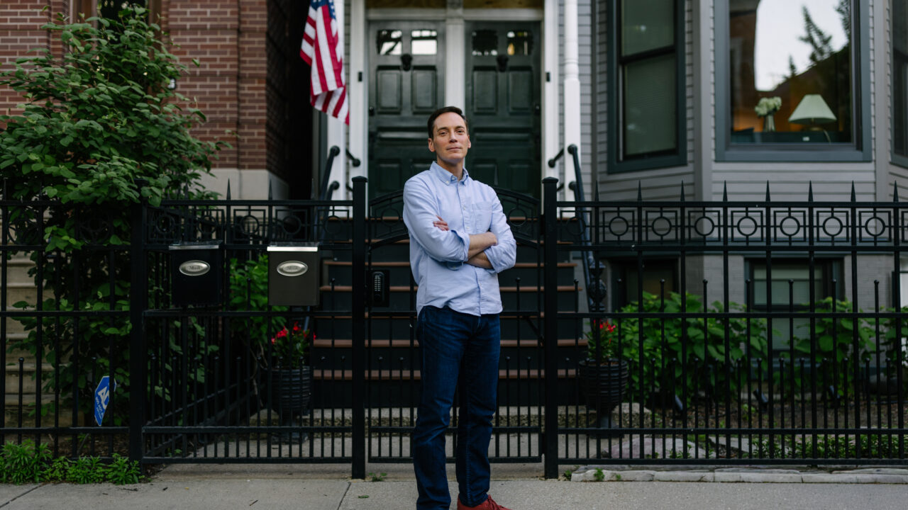 Man with crossed arms standing on sidewalk in front of brick building with green door, decorative black iron fence, and American flag