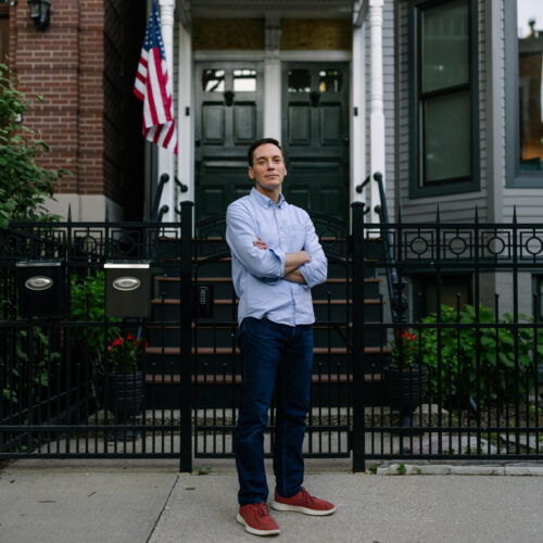 Man with crossed arms standing on sidewalk in front of brick building with green door, decorative black iron fence, and American flag