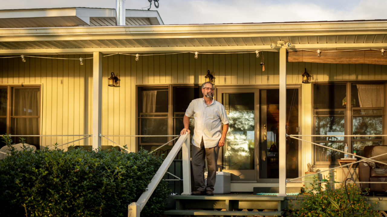Man with glasses in light shirt standing on deck of yellow home with lantern-style lights and green plants