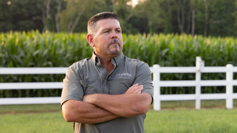 Man with arms crossed standing in front of white fence, cornfield, and trees at Sun Valley Orchards in New Jersey