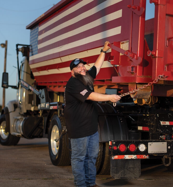 Man in black shirt and cap standing beside red dumpster truck painted with American flag design