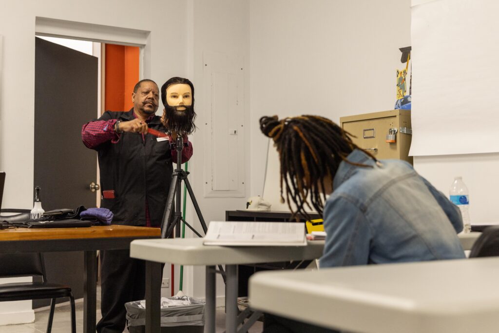 Instructor demonstrating barbering techniques on a mannequin head while a student with locs observes in a classroom